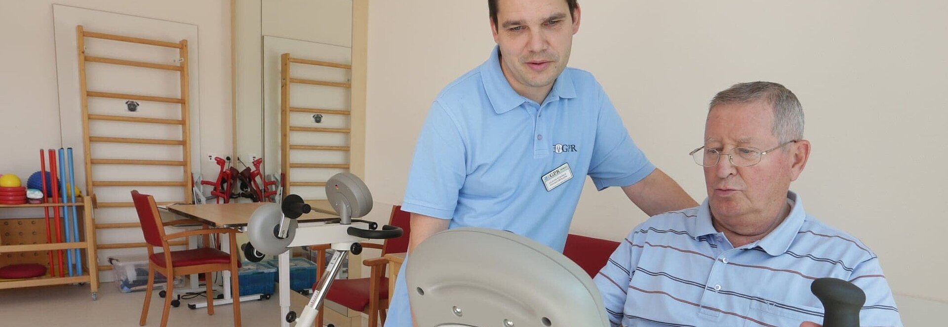 Betagter Patient sitzt auf Trainingsgerät und wird von Physiotherapeut betreut